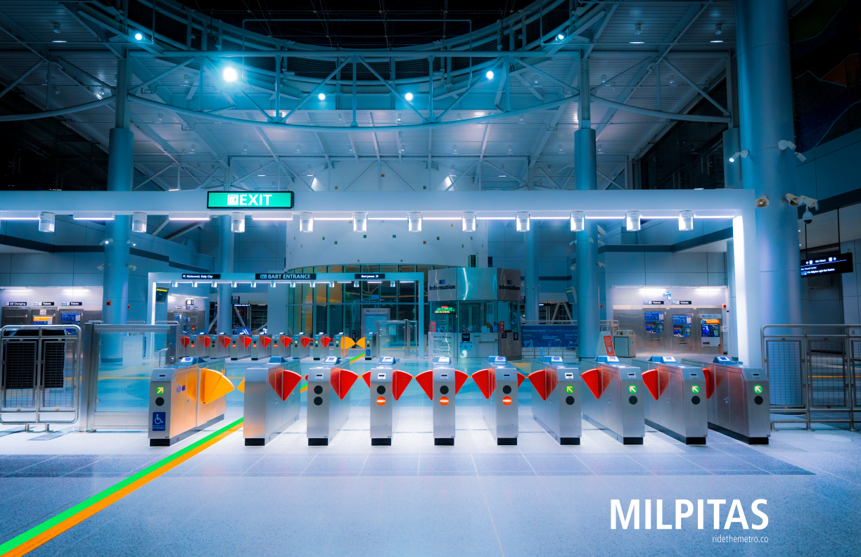 A photo poster that says Milpitas, showing a soft photo of the station lobby at night. The lobby is a large open greyish white space, with several pillars and a large number of metal struts supporting the roof and what appears to be a circular skylight. The room is primarily lit by cool spotlights. In the foreground are the fare gates, over which is a long rectangular arch with a row of spotlights looking down, casting additional light on the gates. There are green and orange lines on the floor that pass-through the accessible fare gate on the left, across the lobby, and out through the accessible gate on the opposite side.